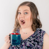 Ashley Doan in front of a white wall, holding up a mug, and opening her mouth in a surprised expression