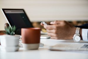 Hands resting on a desk beside an open laptop and plants, holding a phone