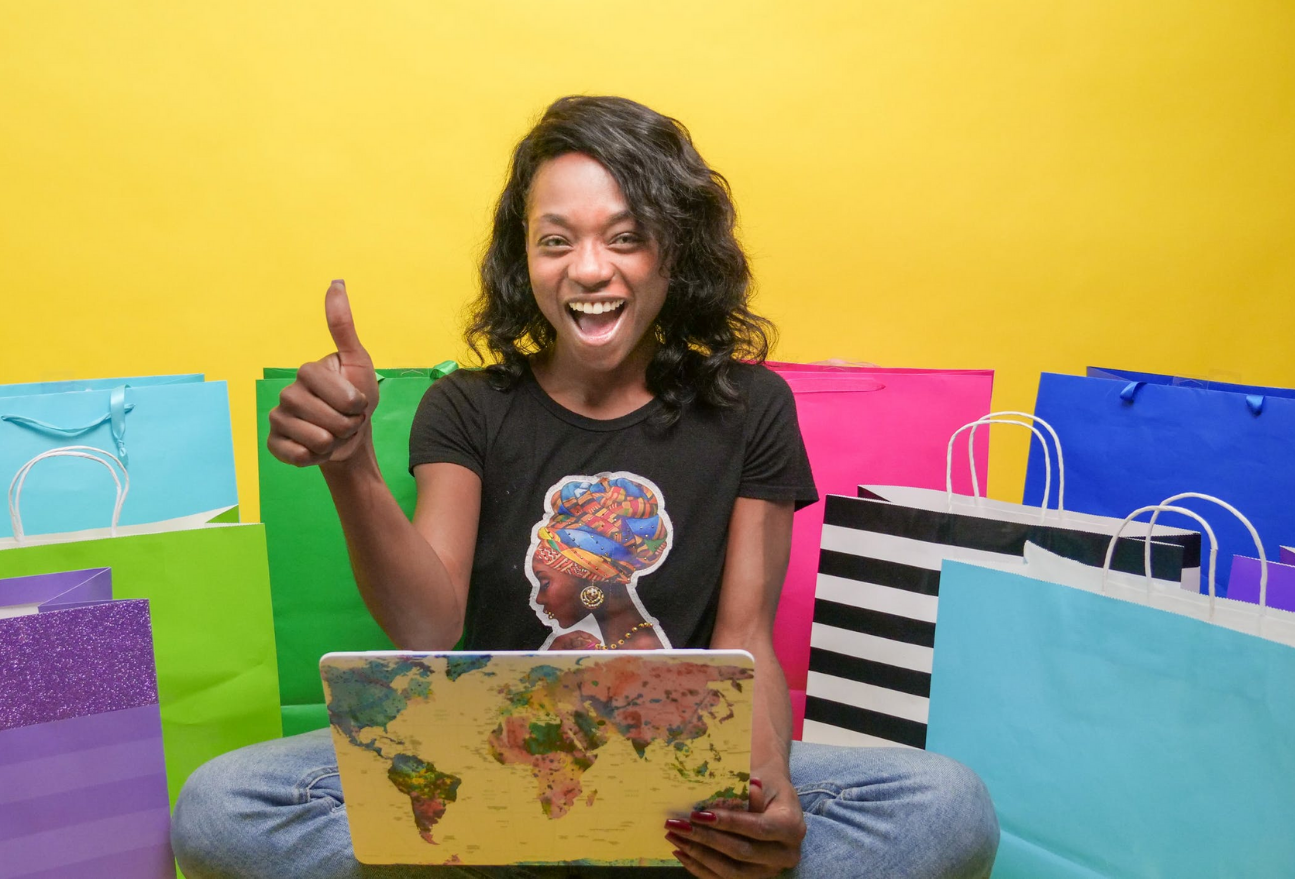 Image of smiling woman using laptop holding up a thumbs up in front of colourful shopping bags