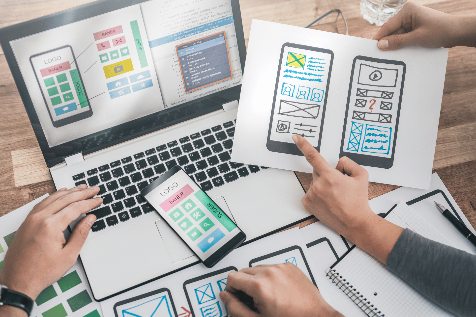 Two people's hands holding papers with website design plans beside an open laptop.