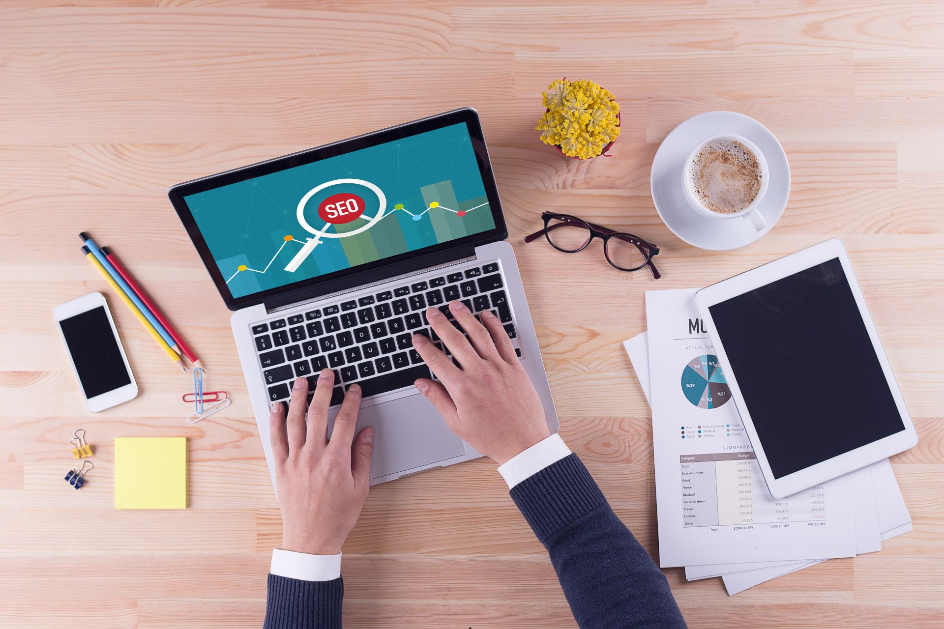 Above angle of hands on desk typing on a laptop with an open screen of a magnifying glass that reads "SEO". Table is covered in office supplies.